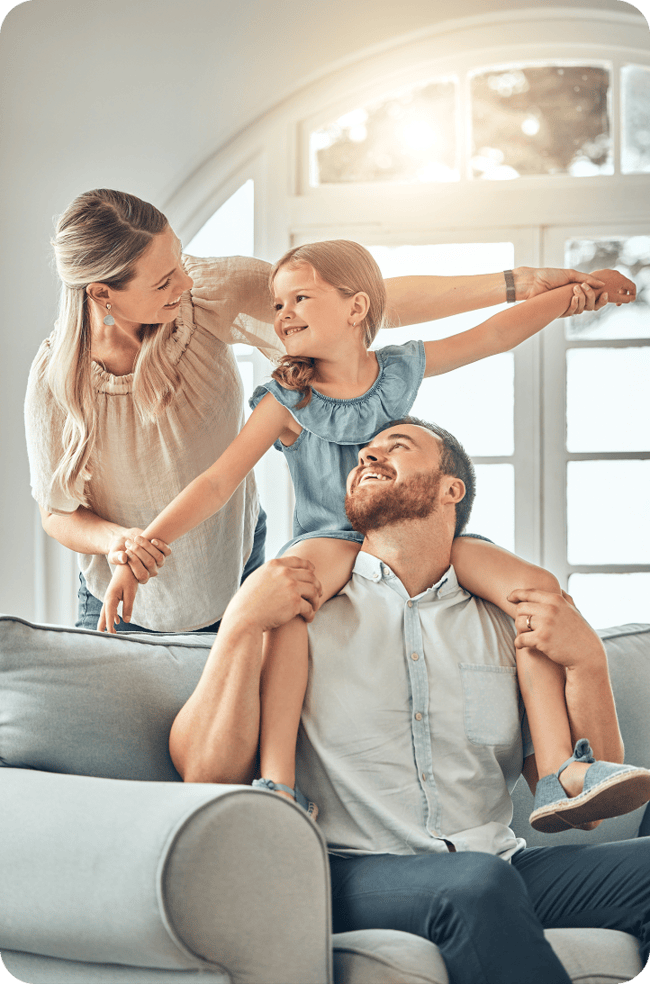 A family of three enjoying time together at home, with a father sitting on a sofa and a daughter on his shoulders, while the mother smiles at them.